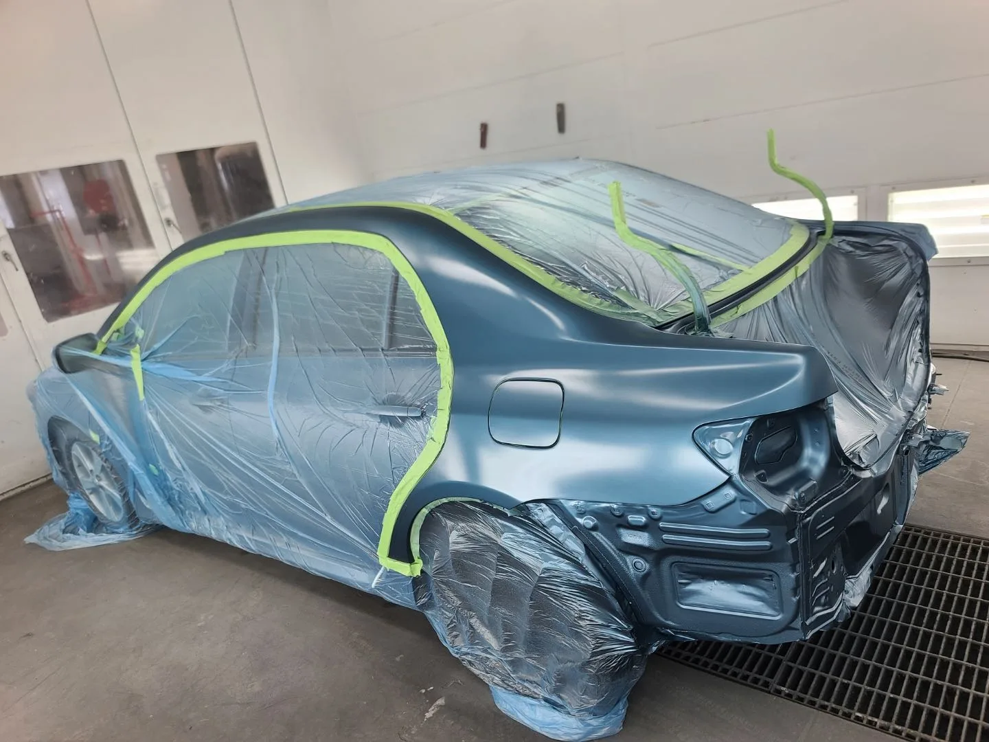 Blue Toyota Corolla masked in the spray booth with primer-coat rear quarter panel visible, mid-repair