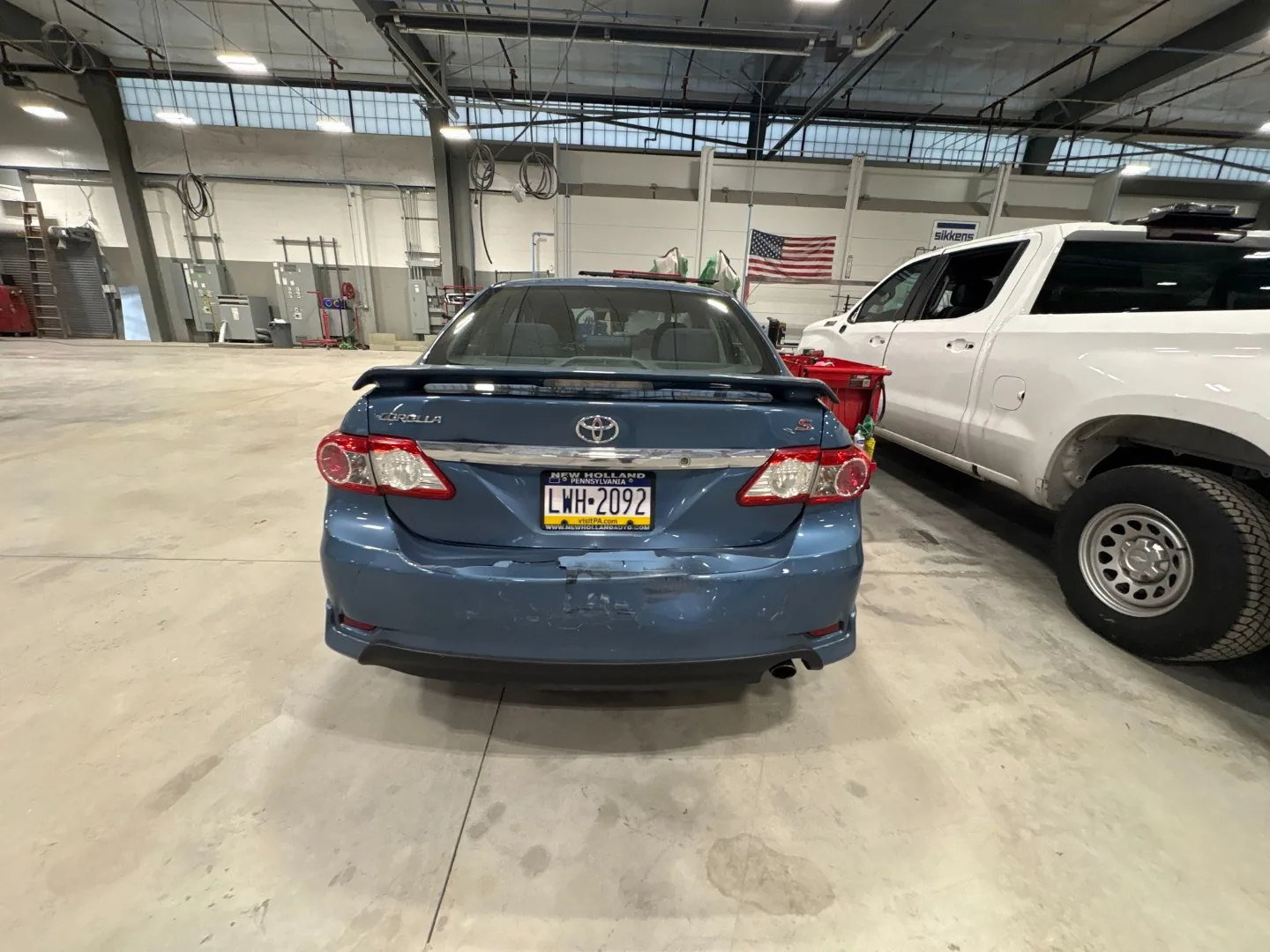 Same blue Toyota Corolla rear view inside the shop showing the original rear bumper and panel damage from the collision