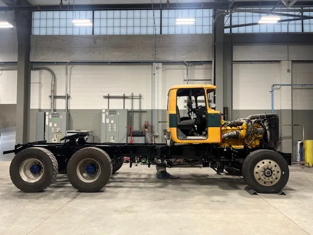 Yellow commercial truck cab on a stripped chassis, full side profile inside the collision repair bay during refurbishment