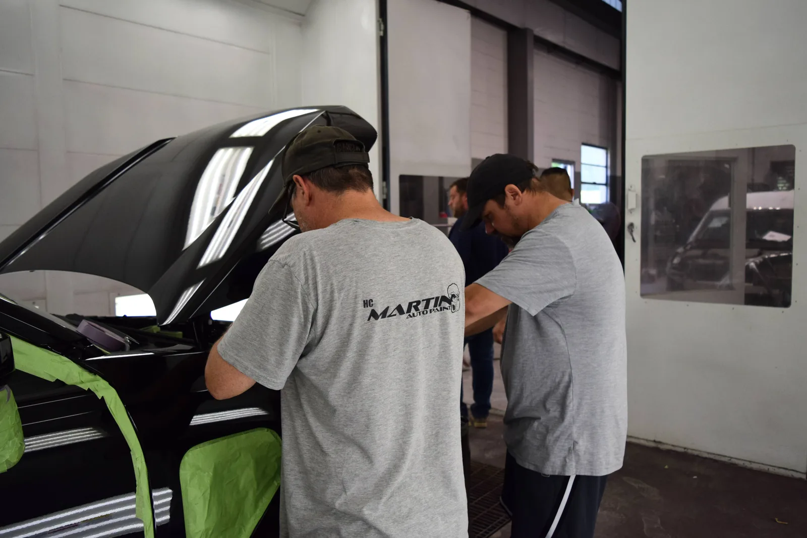 Two Heritage technicians prepping a black hood with green masking in the booth before paint protection work