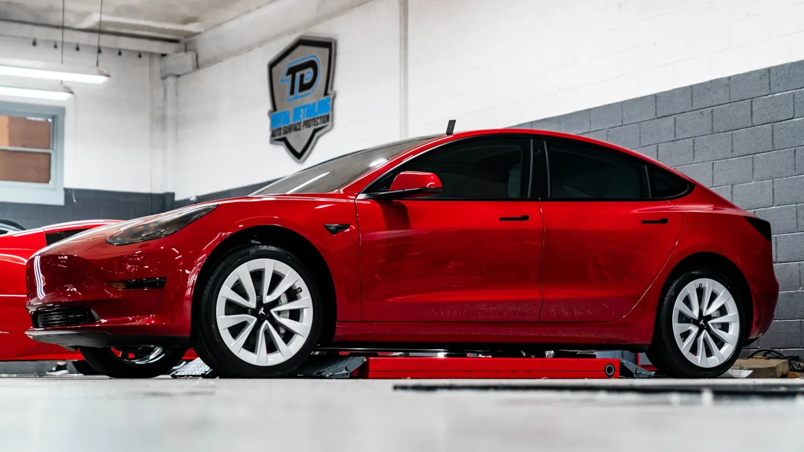 Red Tesla Model 3 side profile under the Total Detailing wall logo, ready for service